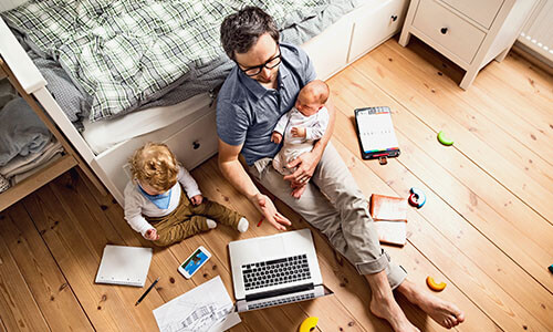 Father holds baby while working on computer and young daughter watches.